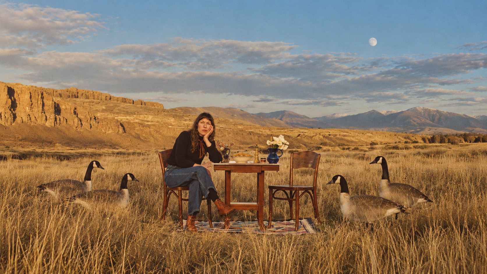 Alela Diane assise à une table dans un vaste paysage doré, entourée d’oies sauvages sous un ciel de fin de journée.