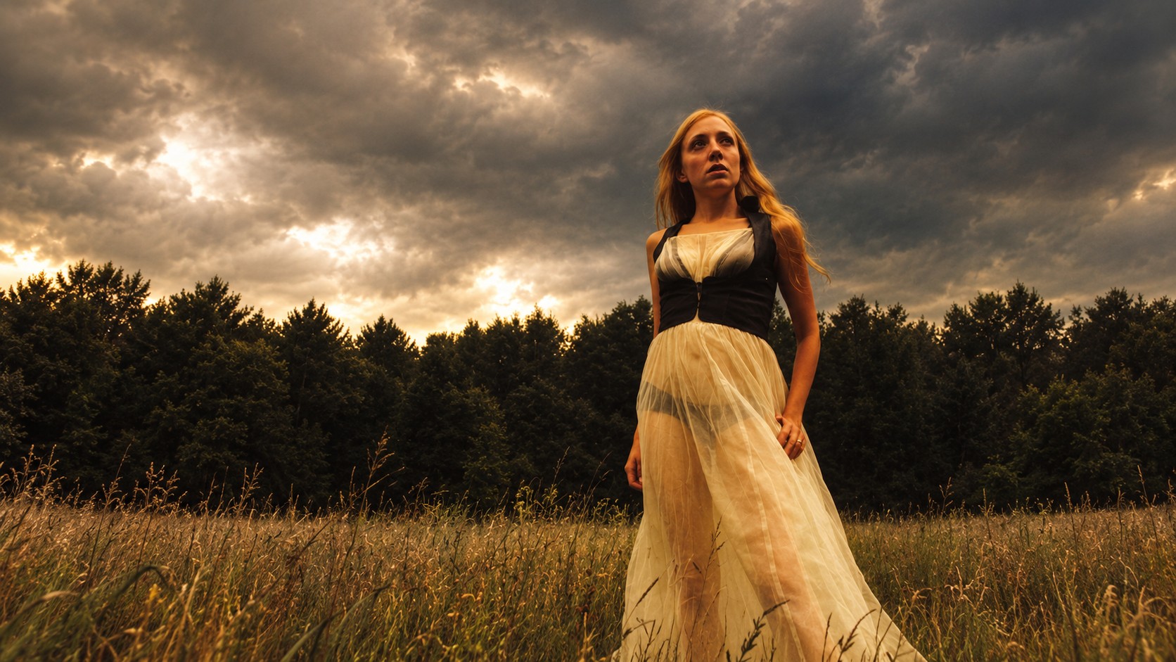 Une femme aux cheveux roux en robe claire dans une prairie, sous un ciel d’orage, devant une forêt sombre.